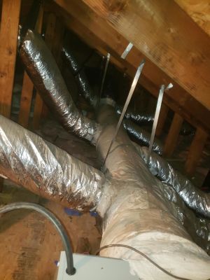 Silver flexible HVAC air ducts suspended by metal straps from wooden rafters in an unfinished attic space.
