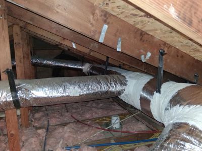 Insulated flexible HVAC ducts suspended from wooden rafters in an attic space with fiberglass insulation.