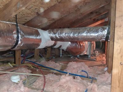 An attic space showing silver insulated air ducts suspended by black straps above pink fiberglass insulation.