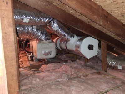 Insulated silver HVAC ducts run through an attic space filled with pink fiberglass insulation between wooden roof rafters.