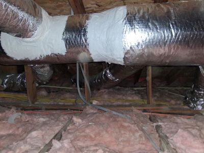 An attic view showing insulated silver ductwork with mastic-sealed joints running across wooden rafters above insulation.