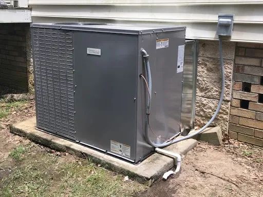 A gray outdoor HVAC unit sits on a concrete pad next to a house with white siding and a stone foundation.