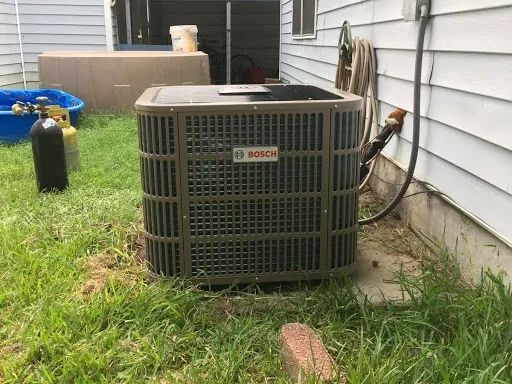 A brown Bosch air conditioning unit sits on a concrete pad next to a house, with a gas cylinder in the background.
