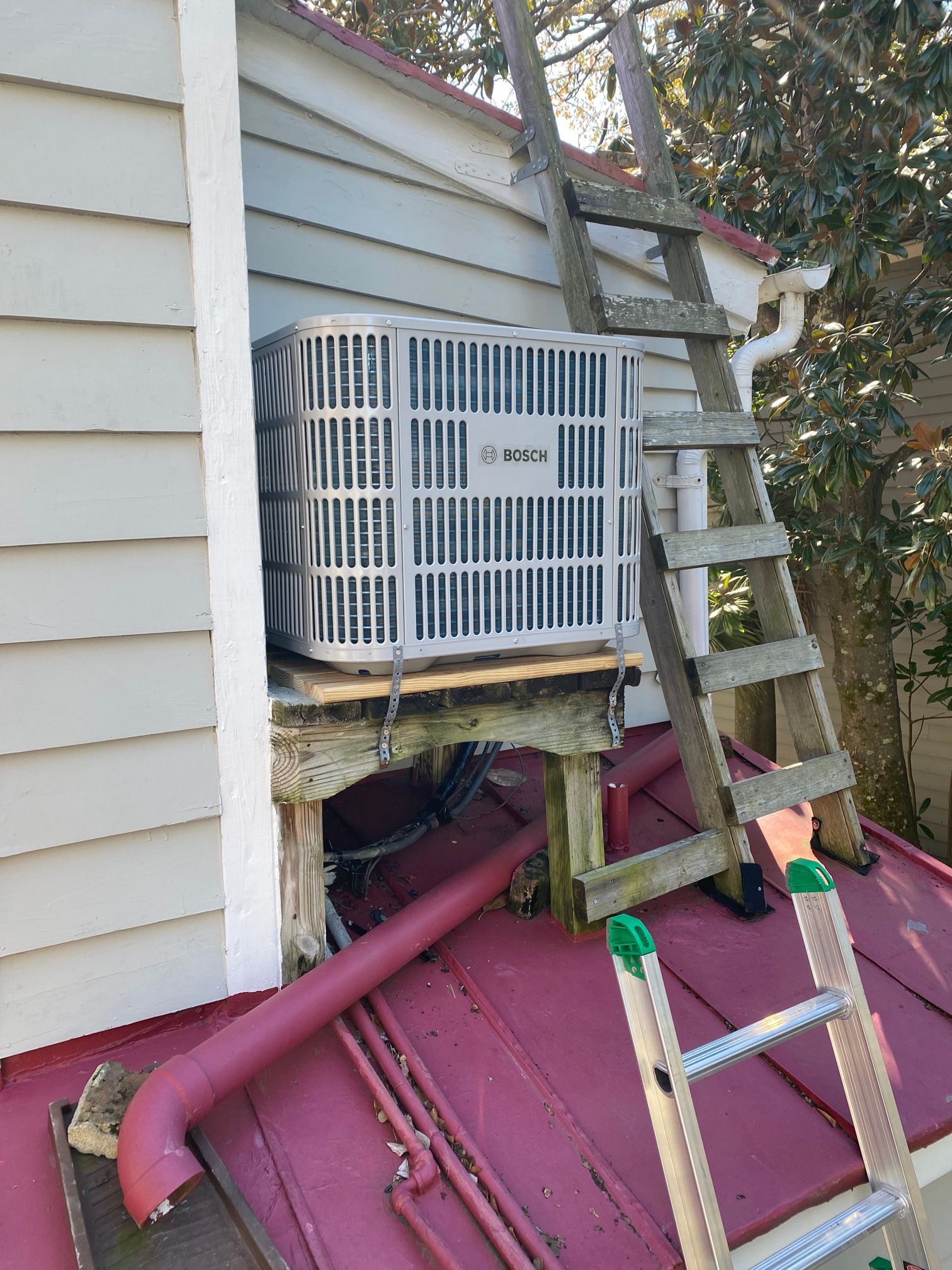 An HVAC condenser unit sits on a wooden platform mounted to the side of a house above a red roof, with ladders nearby.