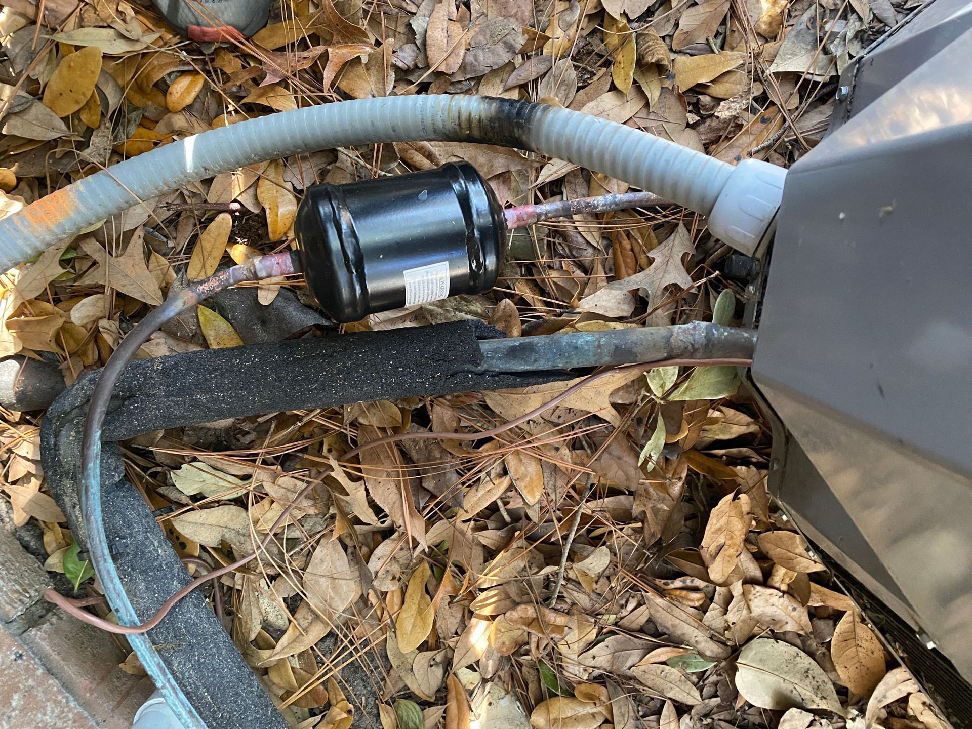A black liquid line filter drier installed on copper tubing connected to an outdoor HVAC unit amidst scattered dry leaves.