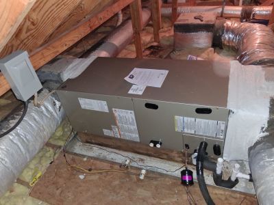 An HVAC air handler unit installed in an attic, connected to insulated silver ductwork and resting on a metal drain pan.