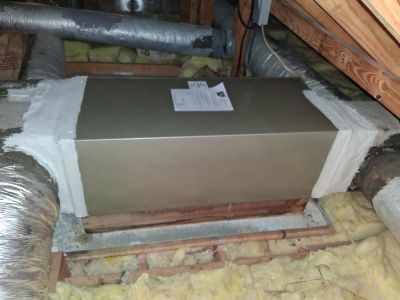 A metal HVAC plenum box sits in an attic, connected to insulated flexible ducts and sealed with white mastic sealant.