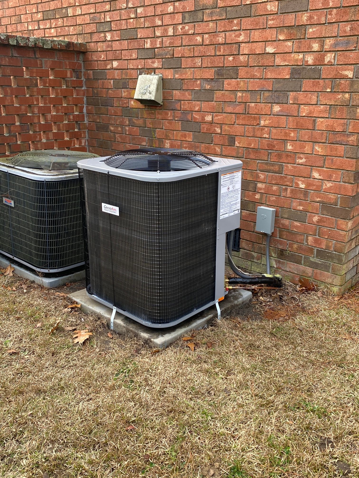 Two grey residential air conditioning units sit on concrete pads against a red brick exterior wall in a grass yard.