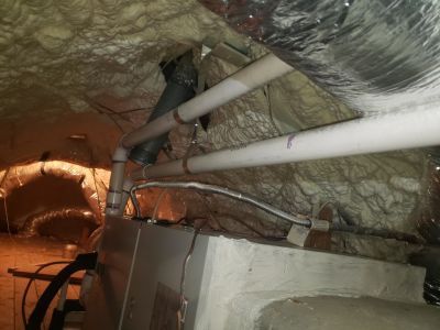 A high-efficiency HVAC unit inside an attic with white PVC plumbing pipes and ductwork against spray-foam insulation.