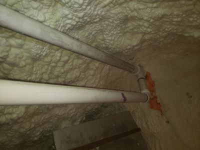 Two white PVC pipes run horizontally along a wall covered in textured, cream-colored spray foam insulation.