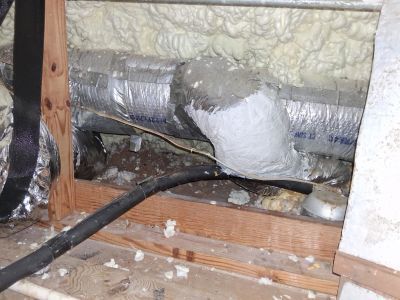 Silver insulated ductwork and a black cable run along wooden joists in an attic with white spray foam insulation.