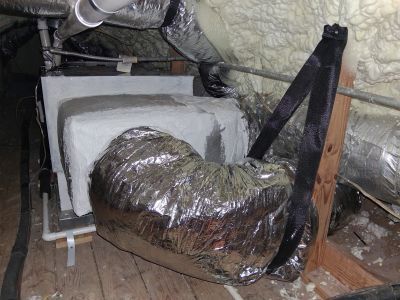 An HVAC air handler unit in an attic, connected to silver insulated flexible ductwork supported by black nylon straps.