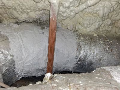 A silver duct sealed with grey mastic paste, crossed by a copper pipe against a backdrop of beige spray foam insulation.