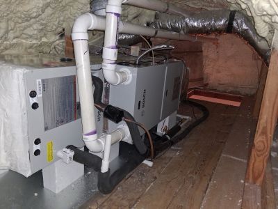 HVAC air handler unit with white PVC piping, located in an attic space with spray foam insulation.