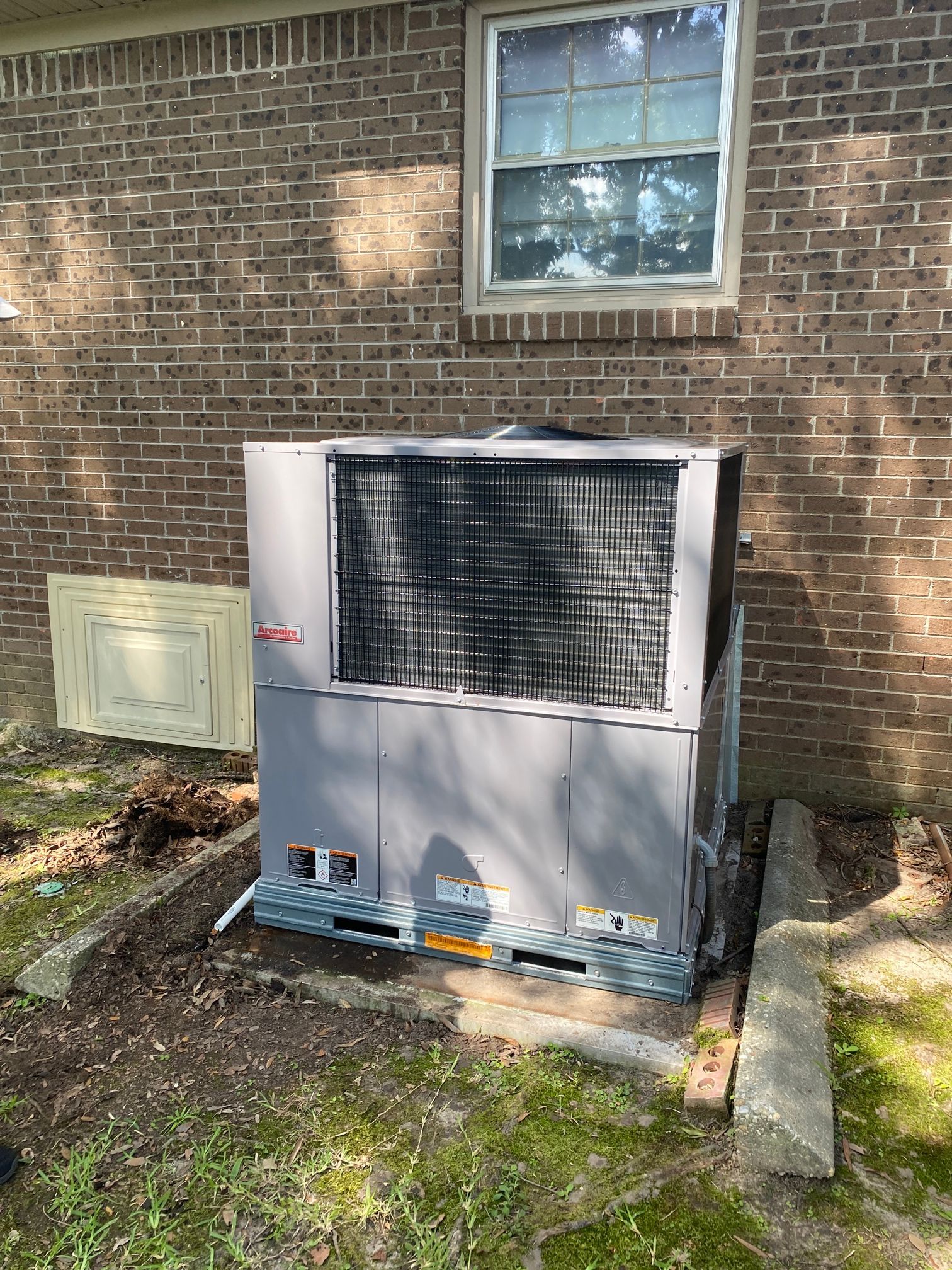 A gray outdoor HVAC unit installed on a concrete pad against a brick exterior wall of a house.
