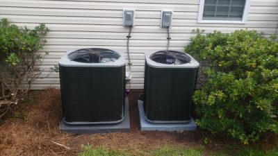 Two outdoor HVAC air conditioning units side-by-side next to a house wall with bushes on either side.