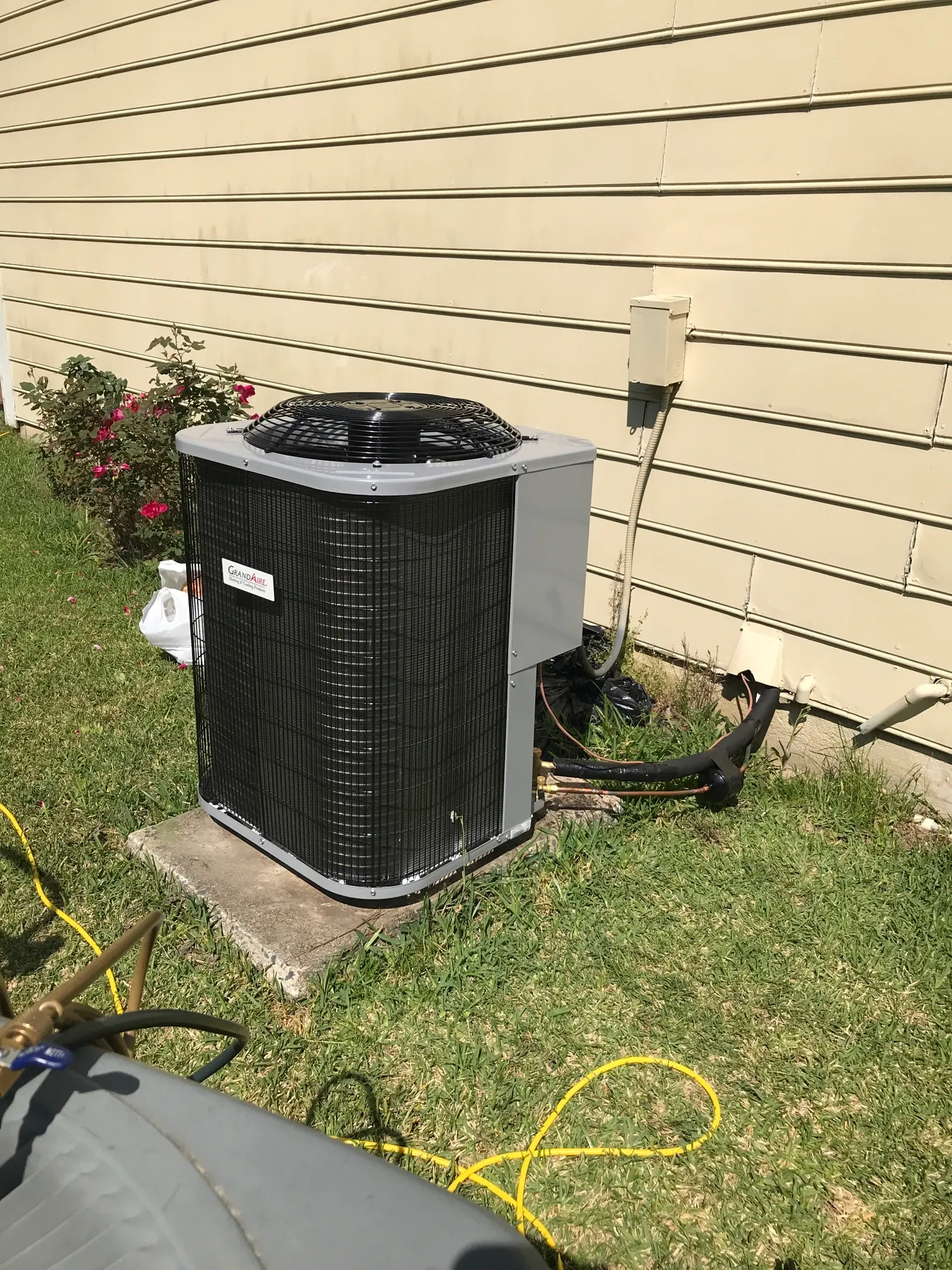 An outdoor air conditioning unit sits on a concrete pad next to a beige-sided house with a small rose bush nearby.