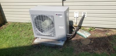 A grey outdoor HVAC unit sits on a concrete pad next to a house with beige vinyl siding and green grass.
