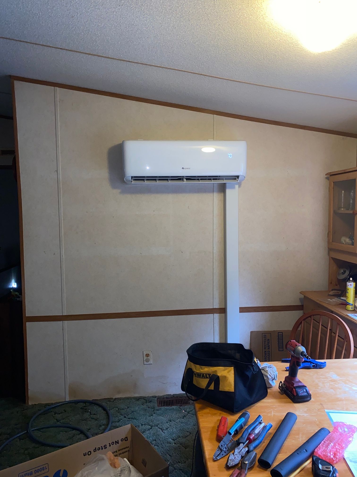 A white ductless mini-split air conditioner mounted on a beige wall, with a white cable cover and tools on a table below.