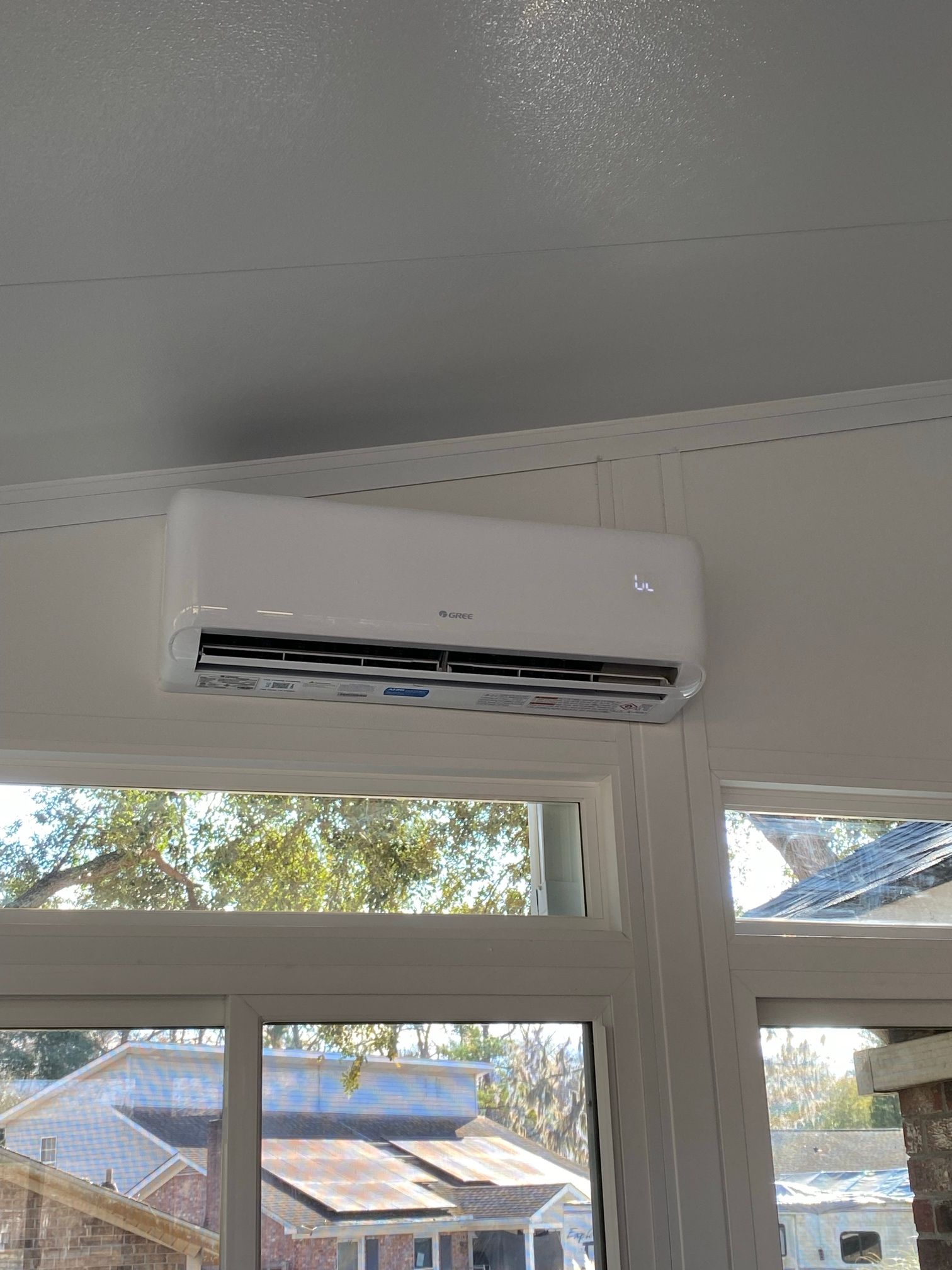 A white, wall-mounted air conditioning unit installed above a window overlooking an outdoor area.