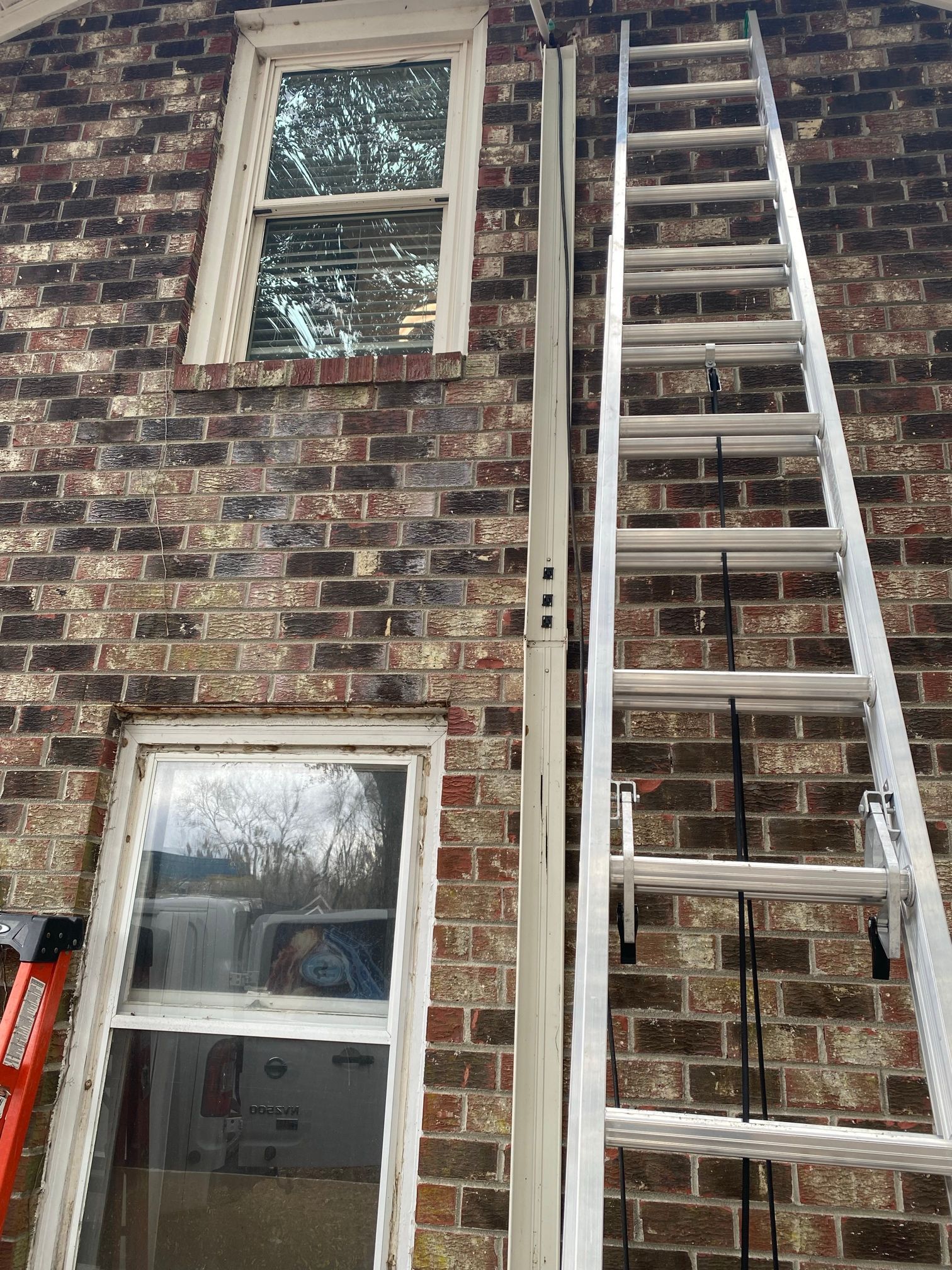 A metal extension ladder leaning against a brick wall between two windows, beside a vertical pipe.