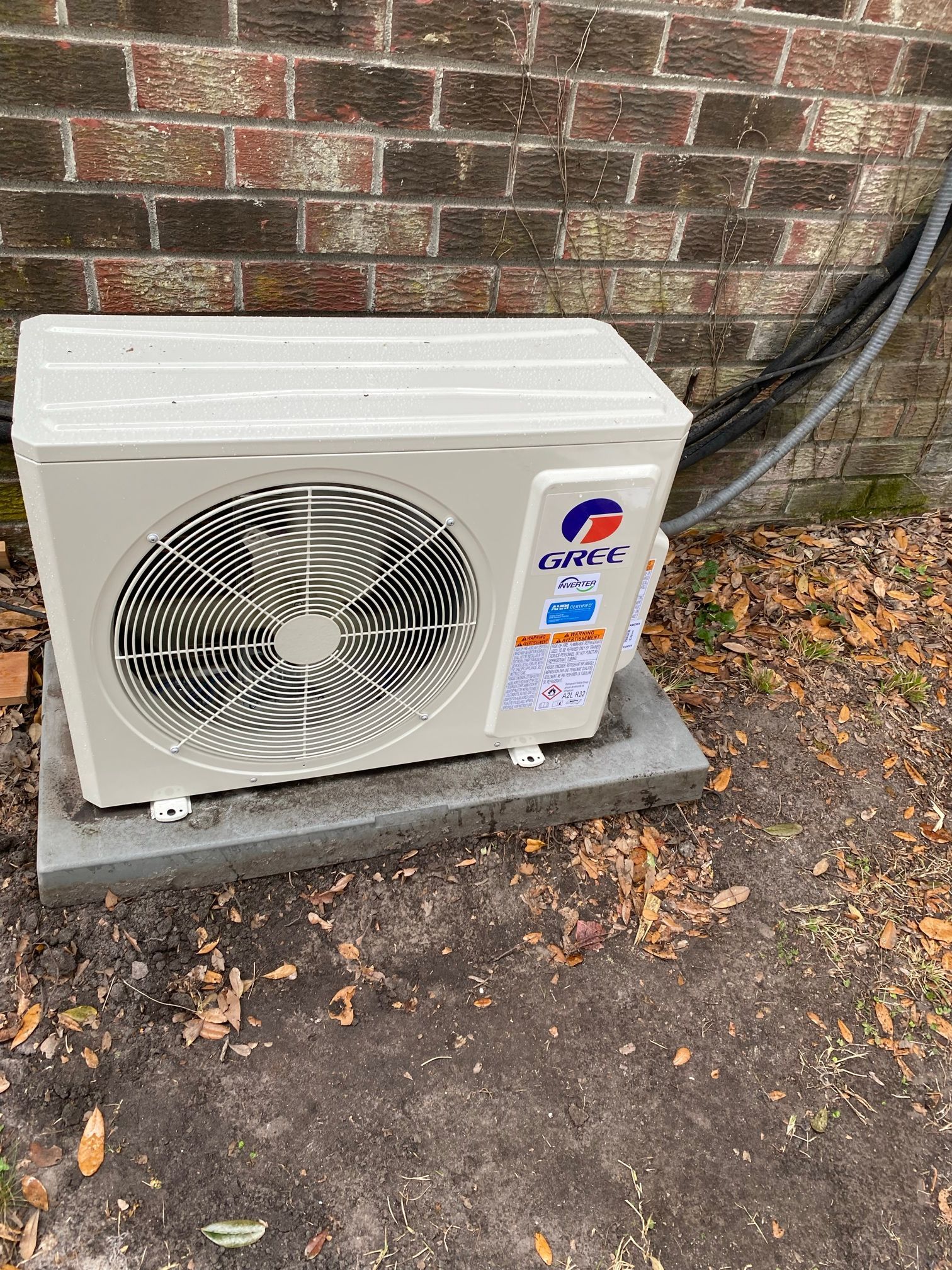 A Gree mini-split outdoor heat pump unit sits on a concrete pad against a brick wall on the ground.