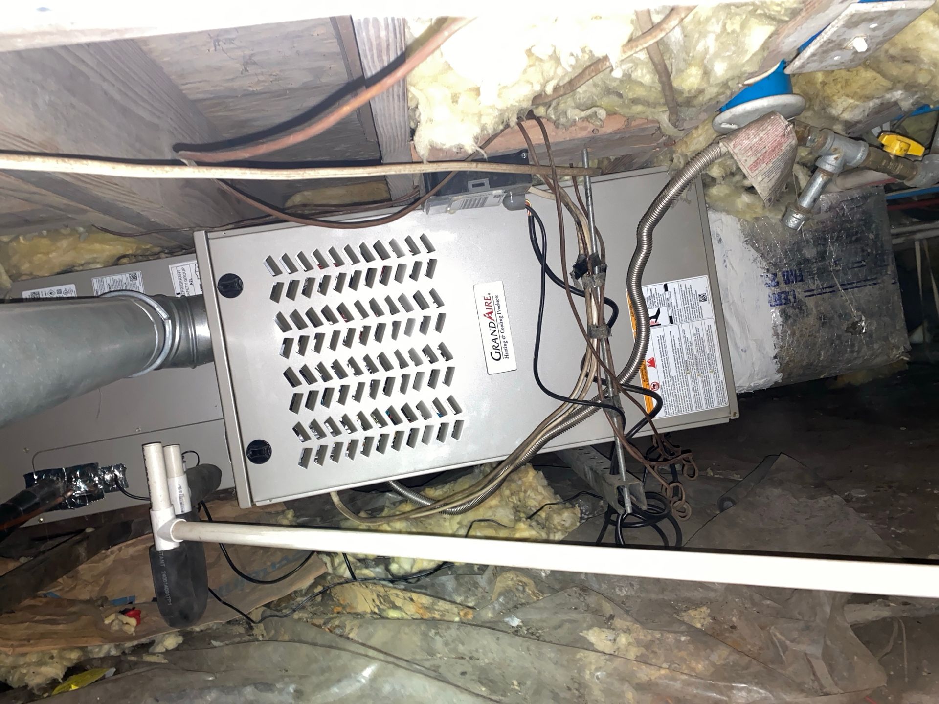 A gray HVAC unit sits in a crawlspace with insulation, exposed wiring, and a white pipe extending into the foreground.