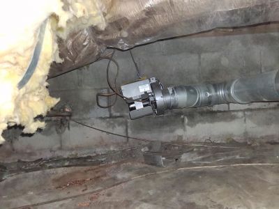 A power venter fan attached to a metal flue pipe installed in a crawlspace under exposed insulation and block walls.