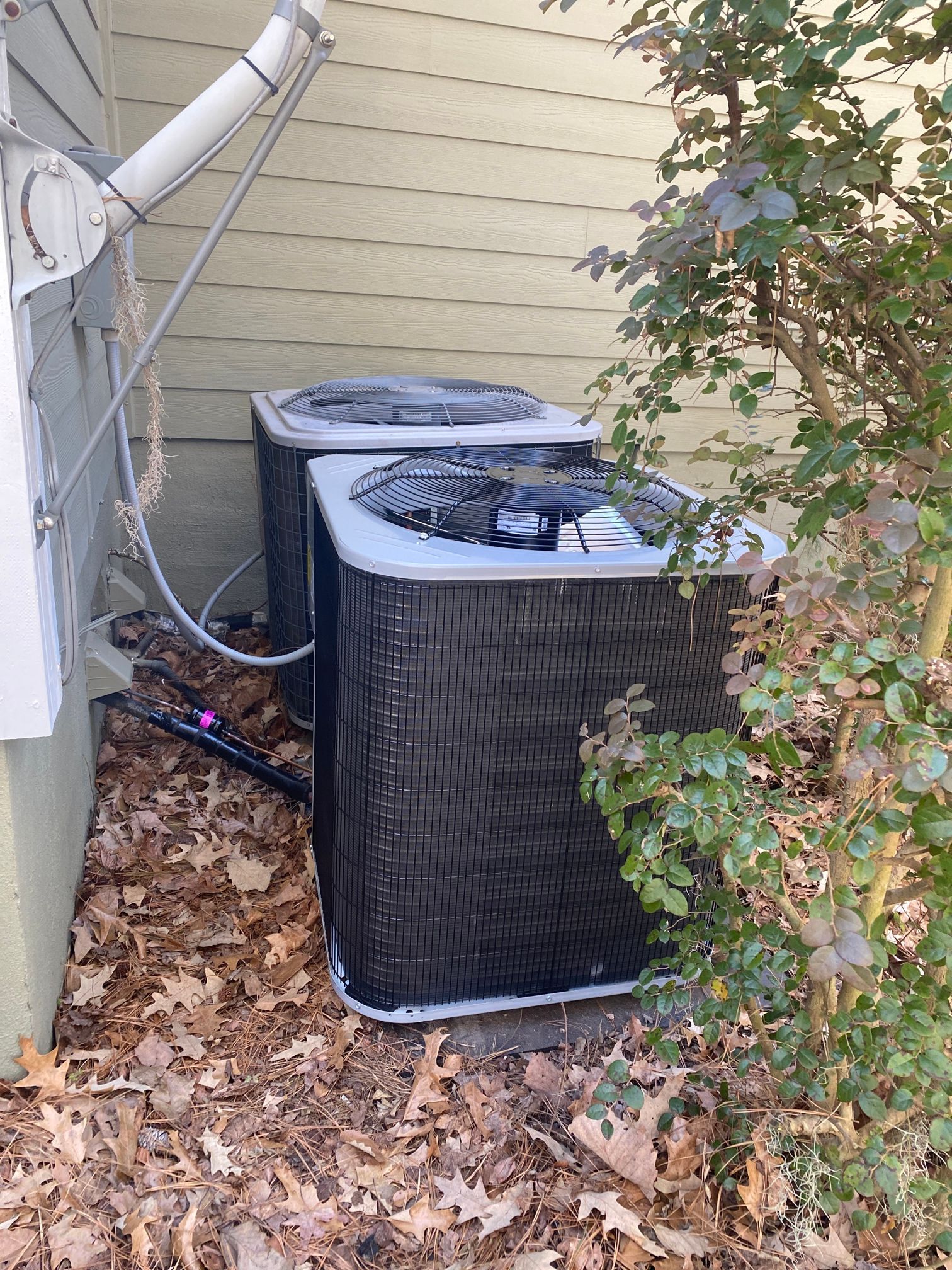 Two black outdoor HVAC condenser units sit on a leaf-covered ground next to a house wall and a leafy shrub.