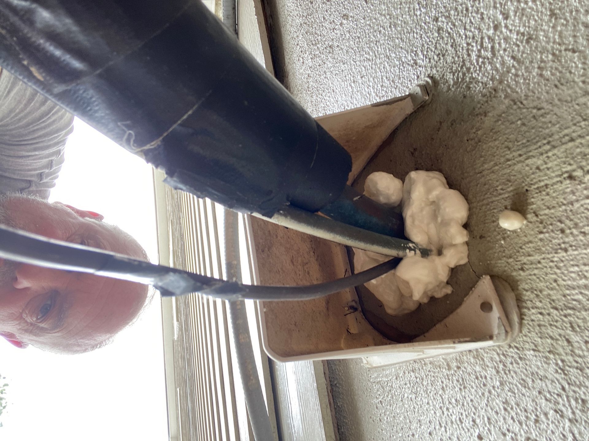 A person using a caulk gun to apply white expanding foam sealant around a cable entering a hole in a concrete wall.