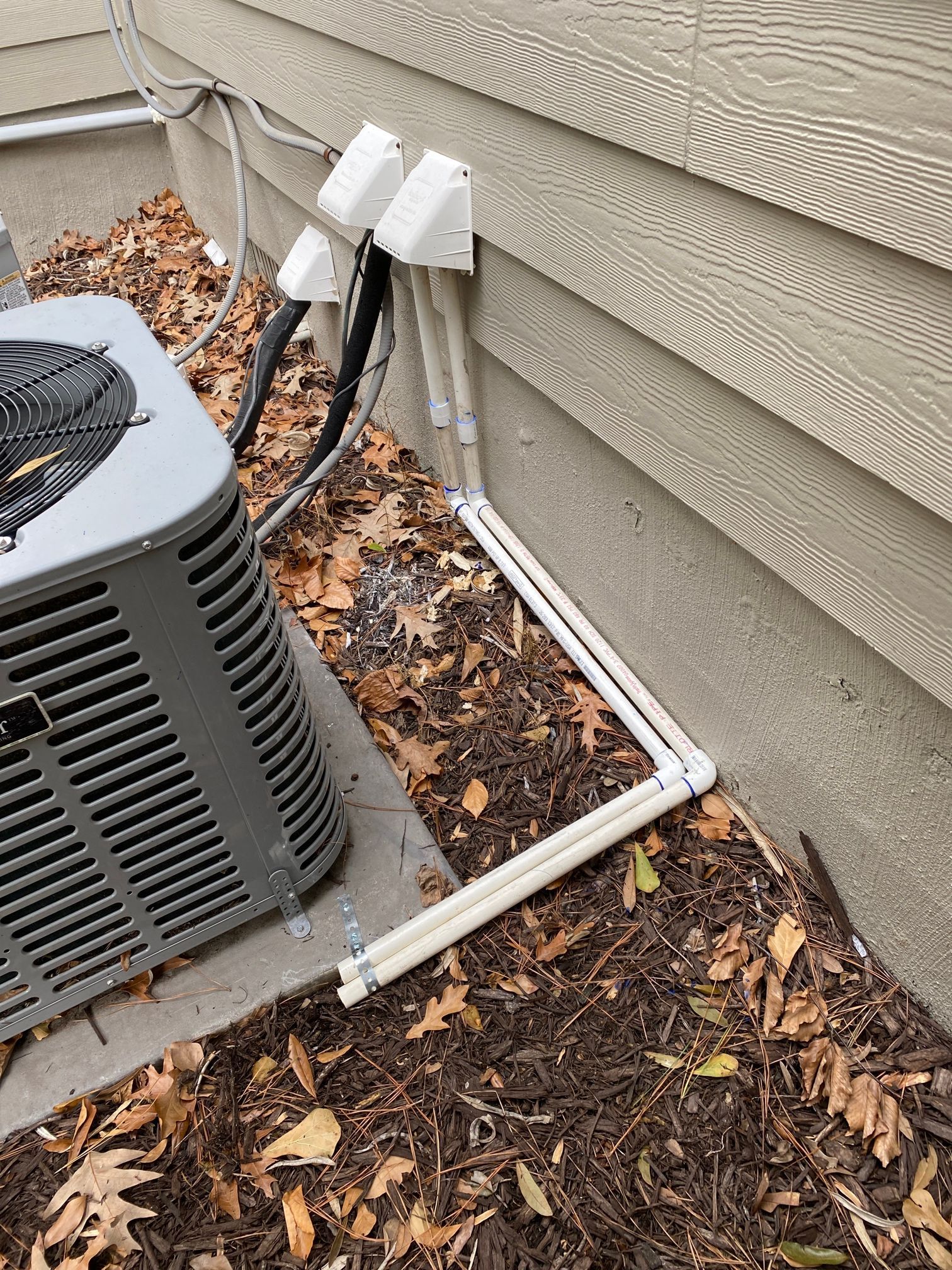 Two white pipes run along a concrete foundation wall next to an outdoor air conditioning unit and tan siding.