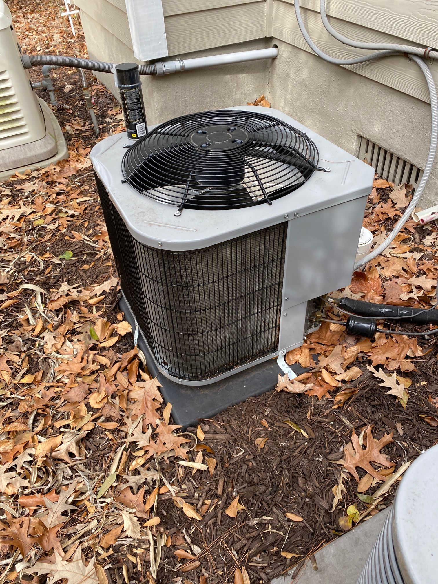 A grey outdoor air conditioning unit sits on a pad in a yard covered with fallen leaves.