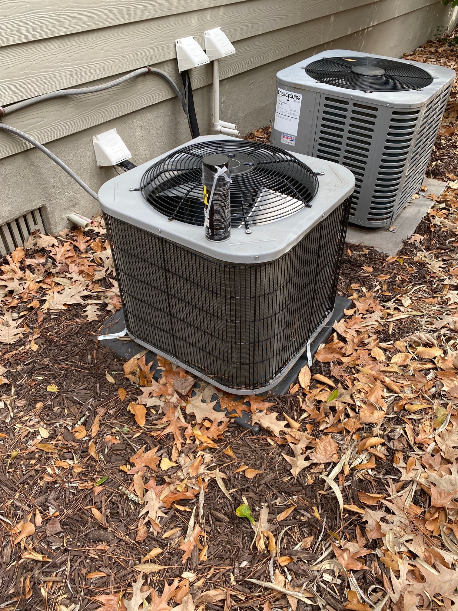 Two gray outdoor HVAC condenser units sit on a ground-level bed of fallen leaves next to a tan siding exterior wall.