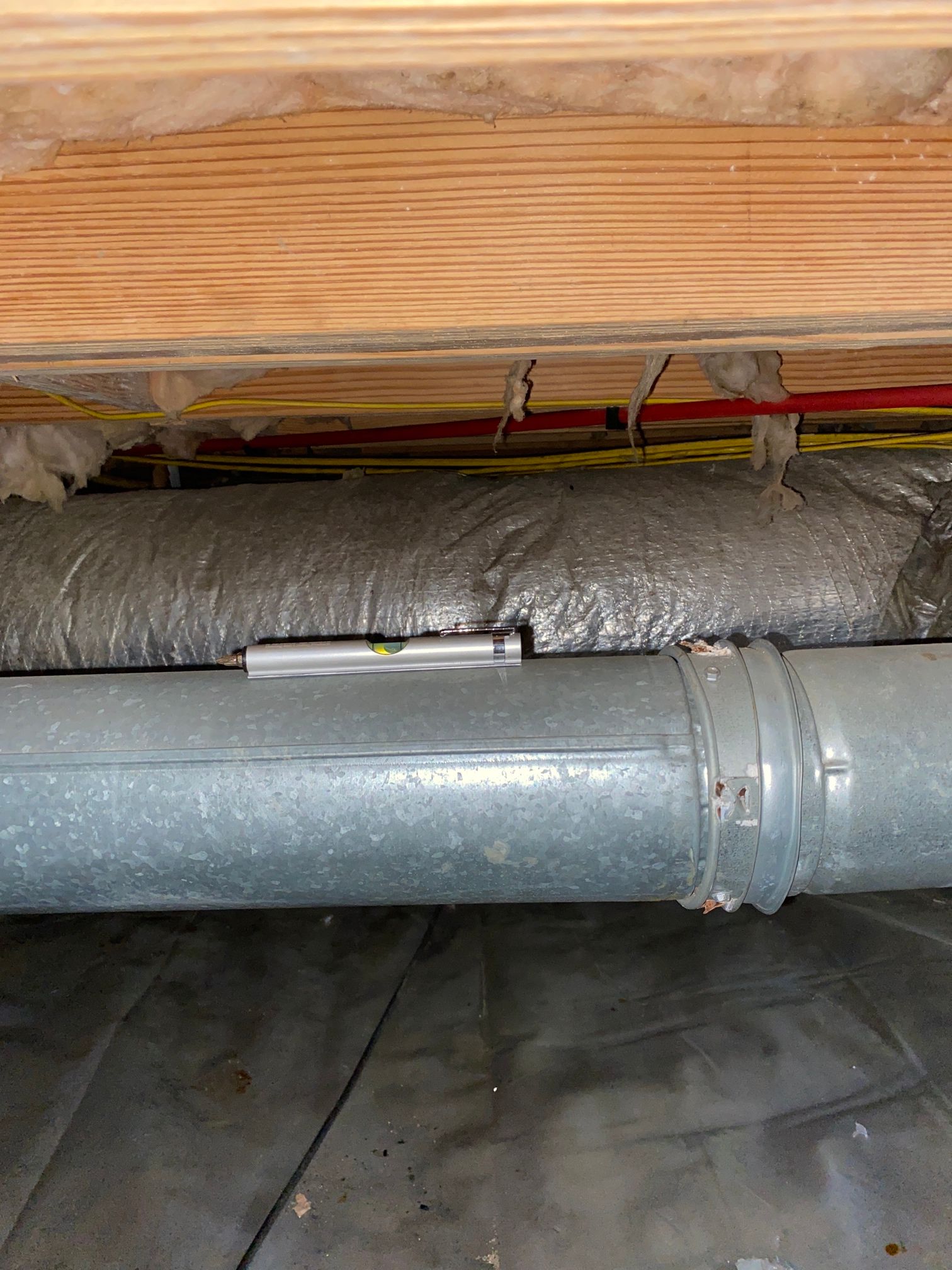 A torpedo level sits on a silver metal HVAC duct in a crawl space under wooden floor joists.