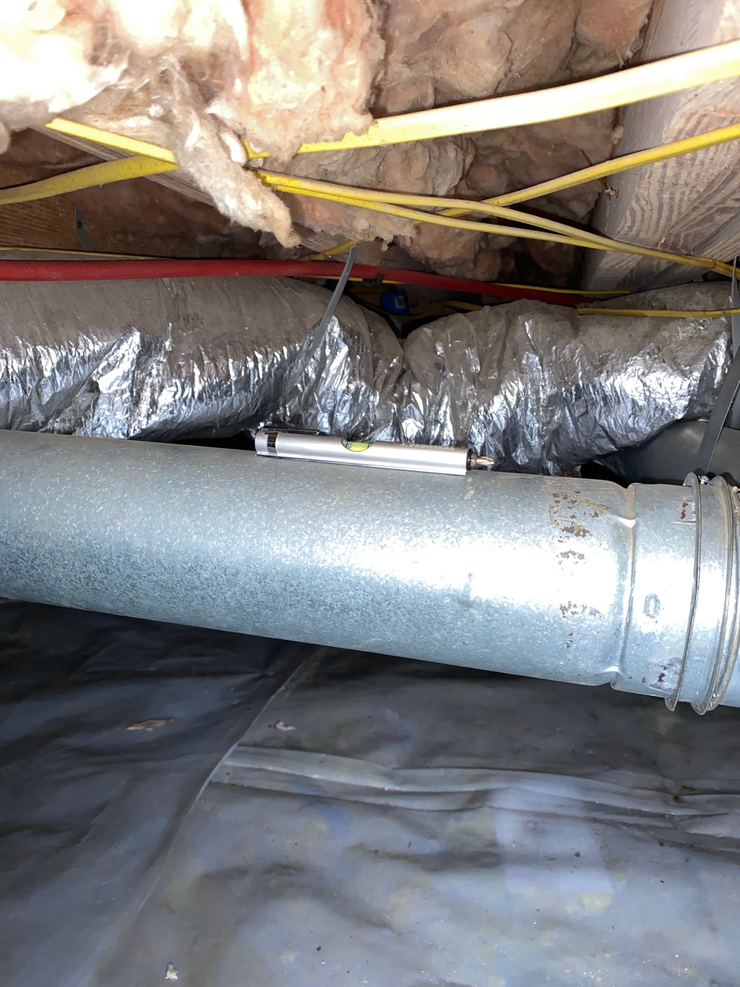 A spirit level sits on a horizontal metal duct in a crawl space with exposed insulation, wiring, and a vapor barrier.