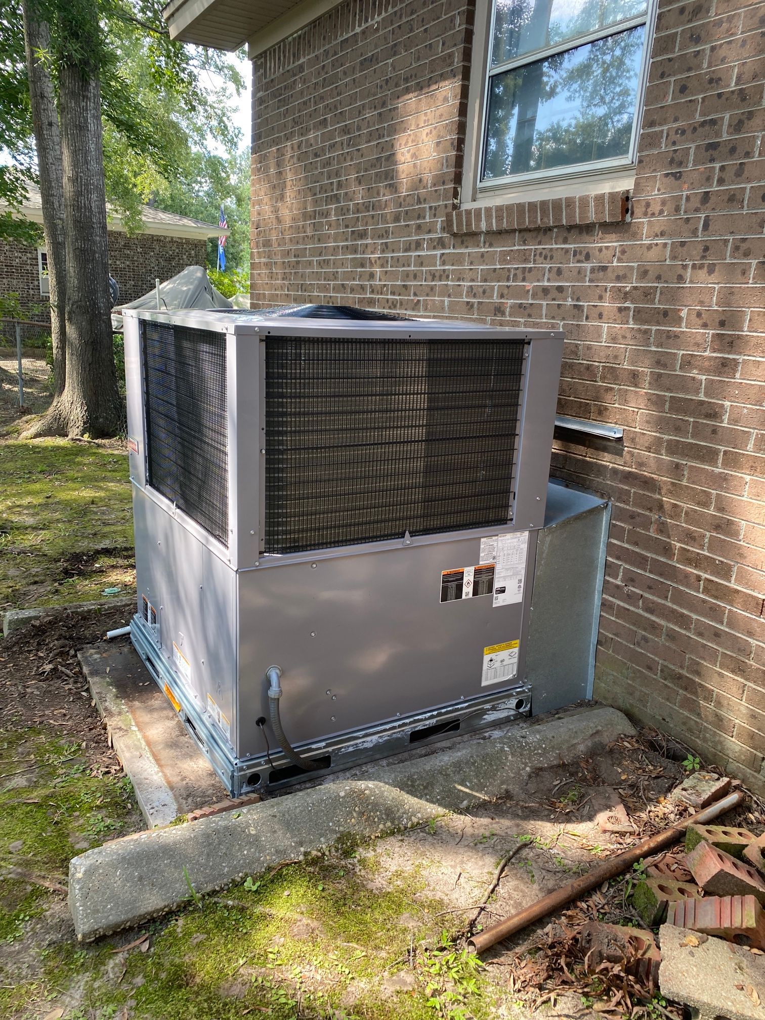 A gray HVAC package unit sits on a concrete pad next to a brick house exterior near trees and yard debris.