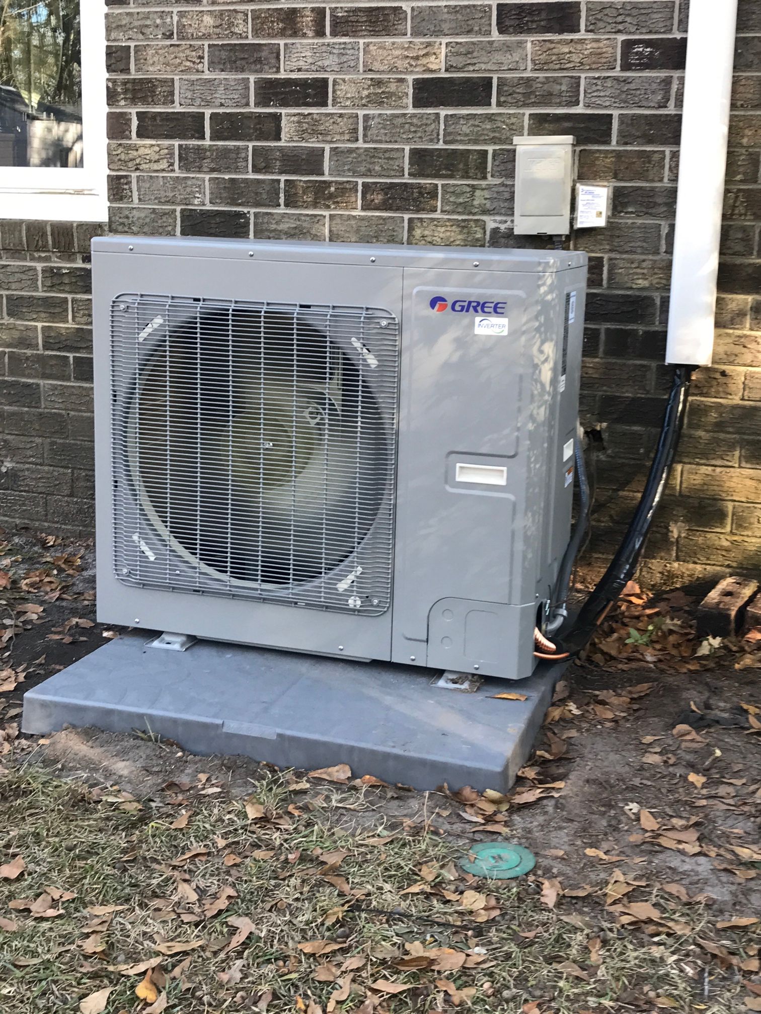 A gray outdoor HVAC unit sits on a concrete pad against a brick wall, with insulated piping leading into the building.