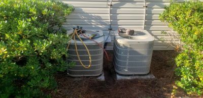 Two grey air conditioning units sit outdoors on a mulch bed next to a house wall, framed by green shrubs.