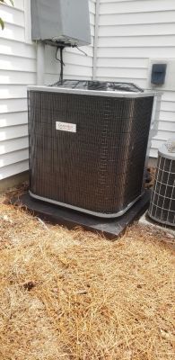An outdoor air conditioning condenser unit placed on a dark base against a white vinyl-sided house.