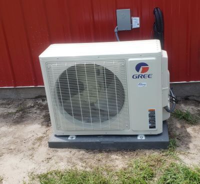 A white Gree mini-split outdoor HVAC unit sits on a dark mounting pad in front of a corrugated red metal wall.