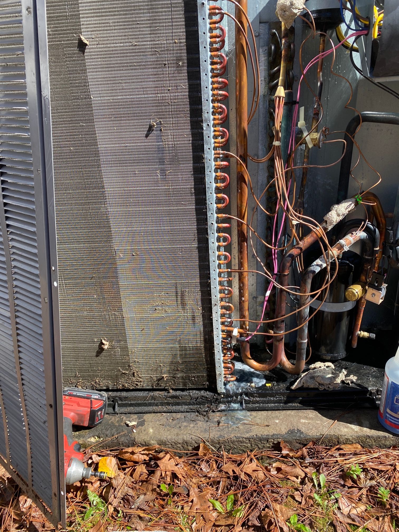 The interior of an outdoor HVAC unit showing copper piping, a metal condenser coil, and exposed wiring on a ground base.