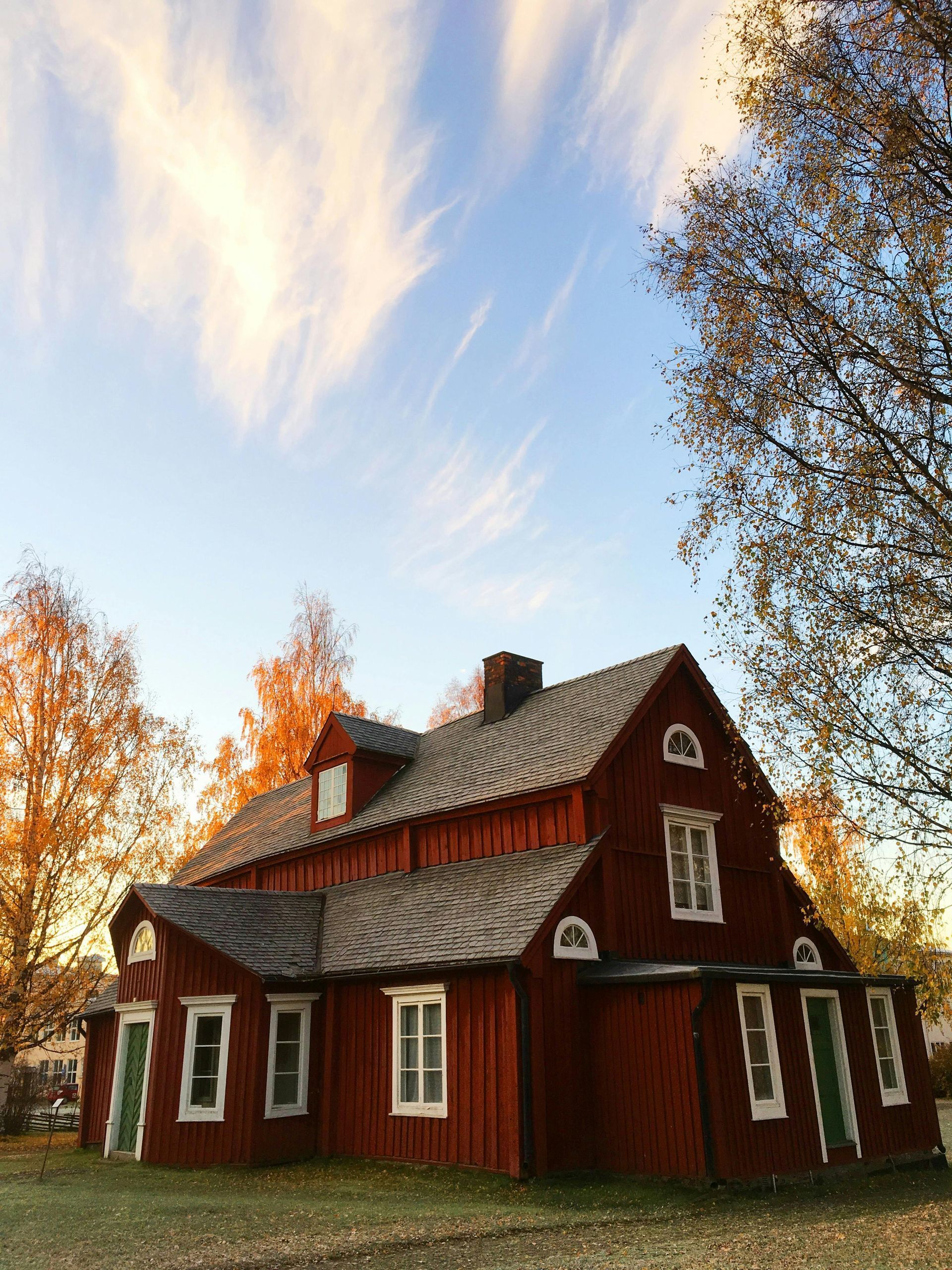 A red wooden house with a grey shingled roof stands surrounded by autumn trees under a blue sky with wispy clouds.