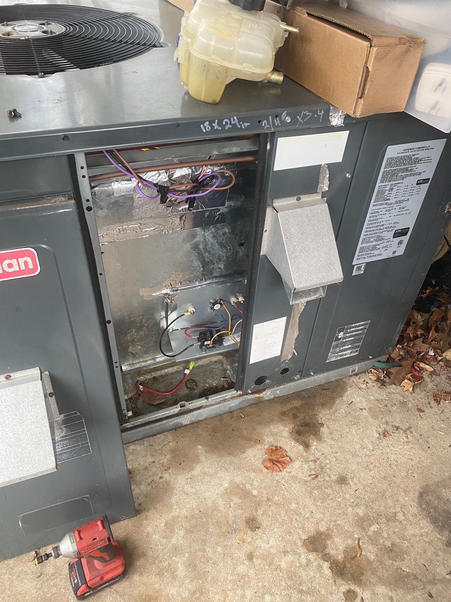 An open outdoor HVAC unit on a concrete surface, with a red power tool on the ground and a plastic jug sitting on top.