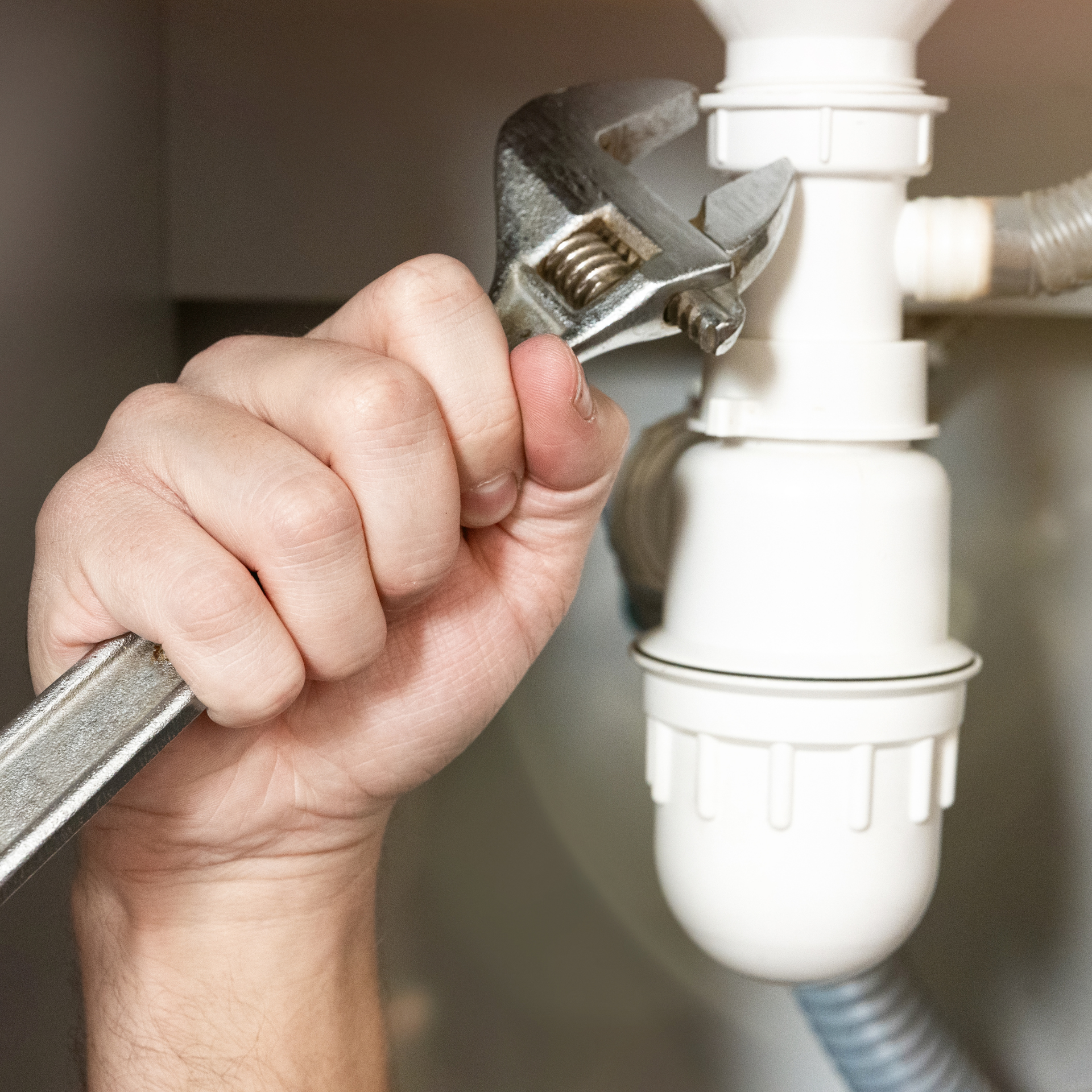 A person's hand uses an adjustable wrench to tighten a white plastic sink drainage pipe.