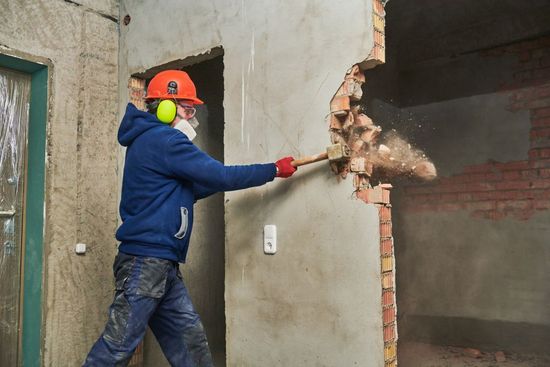 Person in safety gear demolishing a brick wall with a sledgehammer. Debris is flying.