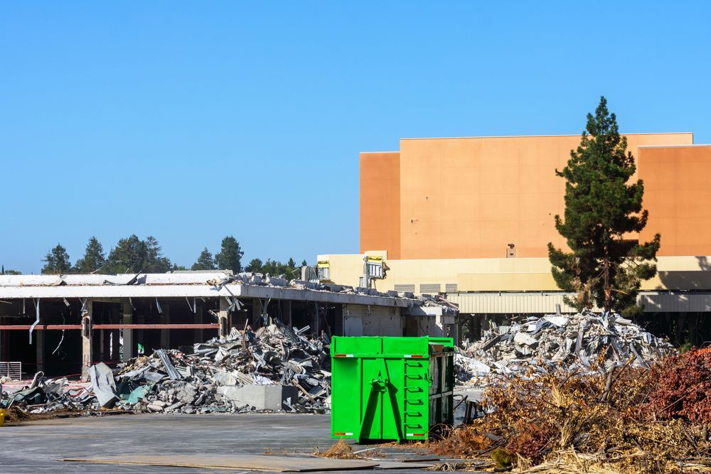 Building demolition with debris pile and green dumpster against blue sky.