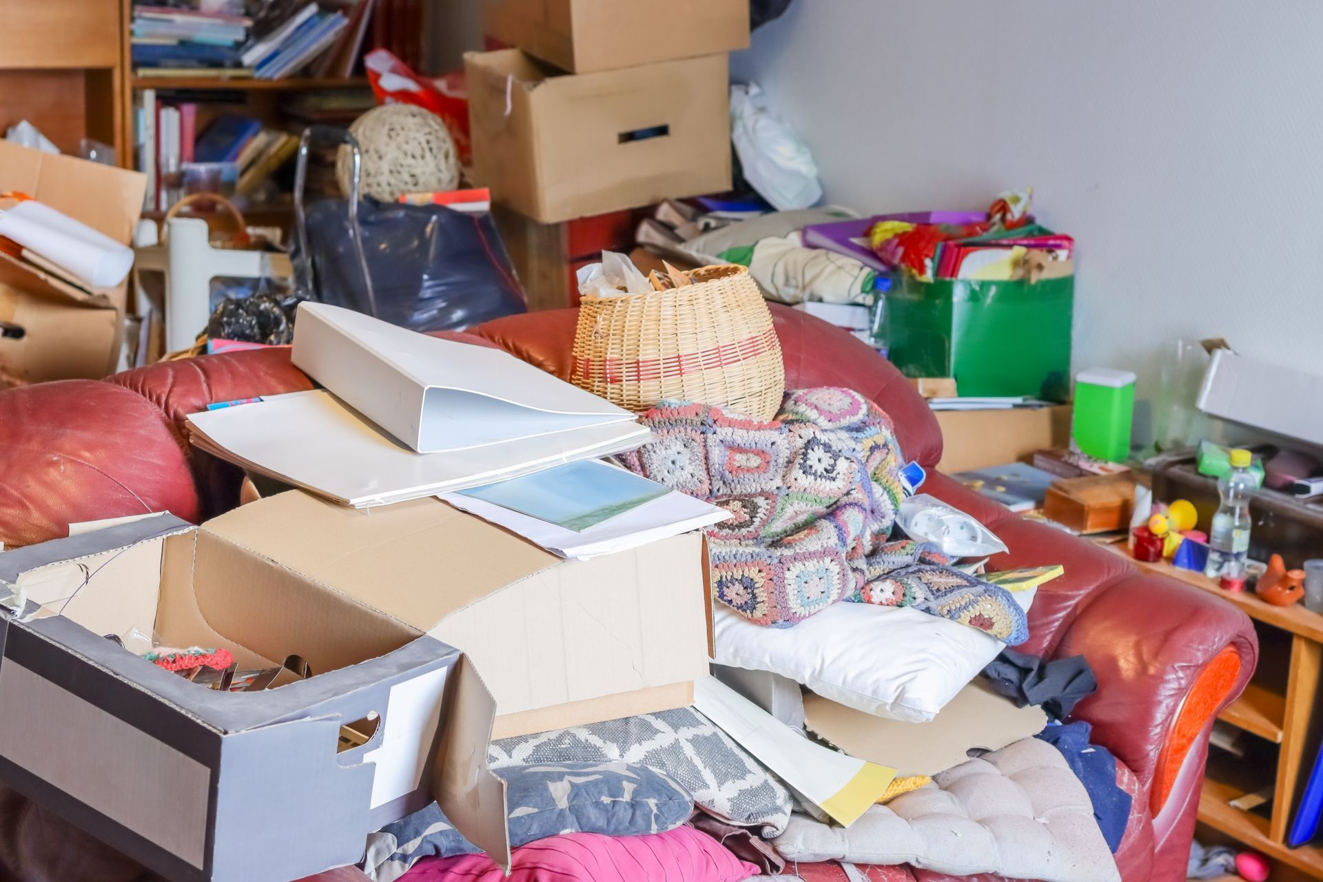 A cluttered red sofa covered with open cardboard boxes, loose papers, and miscellaneous household items in a room.