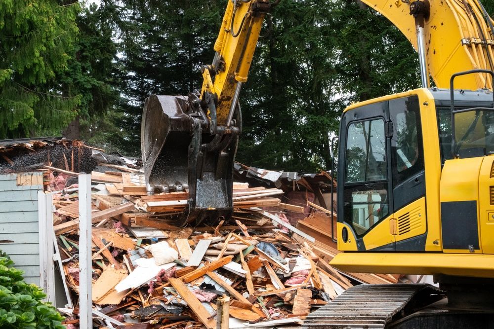 Yellow excavator demolishes a house, surrounded by wooden debris.