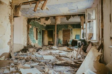 Interior room in disrepair, debris on floor, walls torn apart, natural light through window.