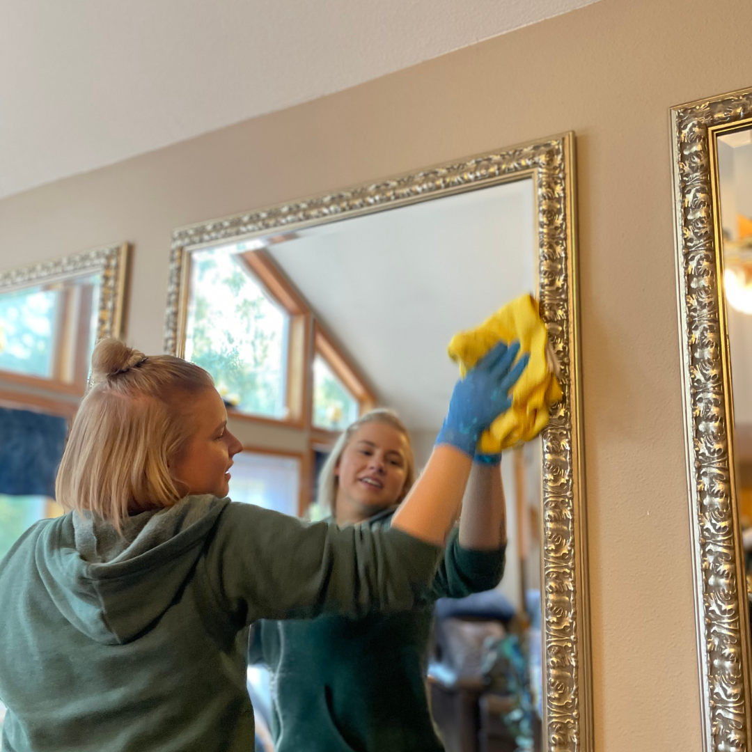 Woman cleaning a large mirror with a yellow cloth, wearing a green hoodie and blue gloves.